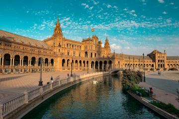 A wide-angle view of the Plaza de España in Seville, featuring its distinctive semi-circular architecture, central fountain, and canal under a blue sky.