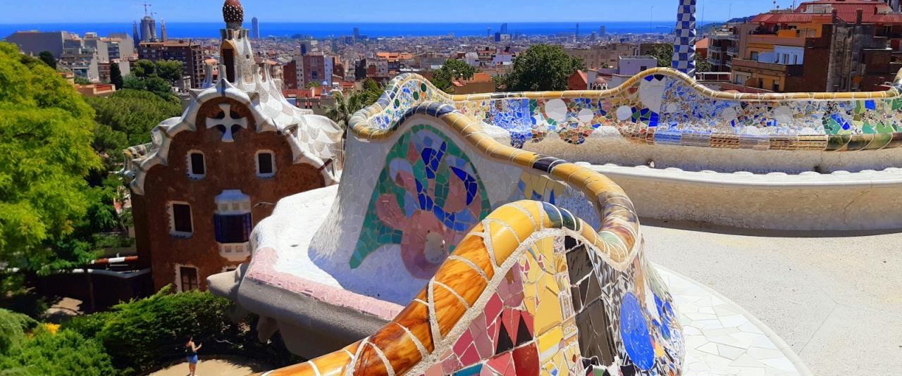 A view of the serpentine mosaic bench and the Dragon Staircase in Park Güell, with the iconic tiled tower and Barcelona skyline in the background.