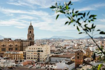 A panoramic view of a dense urban area featuring a prominent cathedral with a tall bell tower in the foreground and mountains in the distance.