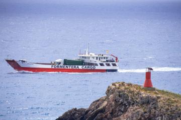 A red and white cargo ferry named "FORMENTERA CARGO" sailing on the open sea, passing a small rocky islet with a red lighthouse in the foreground.