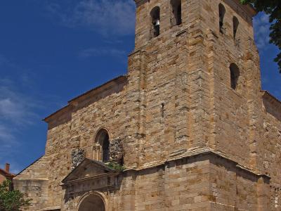 A low-angle shot of a large, historic stone church with a prominent square bell tower featuring arched openings and a tiled roof, set against a blue sky with clouds.