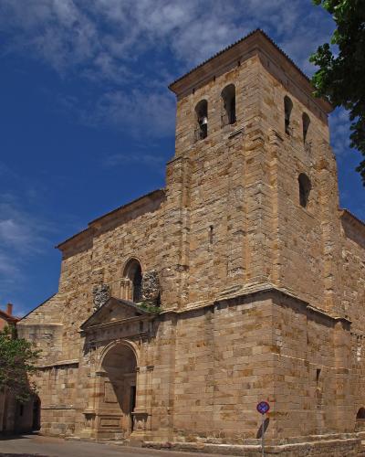 A low-angle shot of a large, historic stone church with a prominent square bell tower featuring arched openings and a tiled roof, set against a blue sky with clouds.