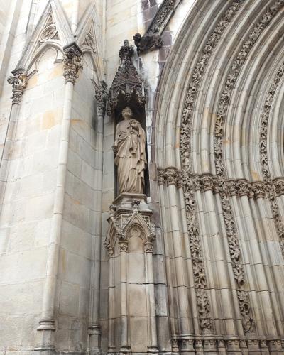 A close-up view of Gothic stone architecture featuring a statue in a niche, a gargoyle, and an ornate arched doorway.