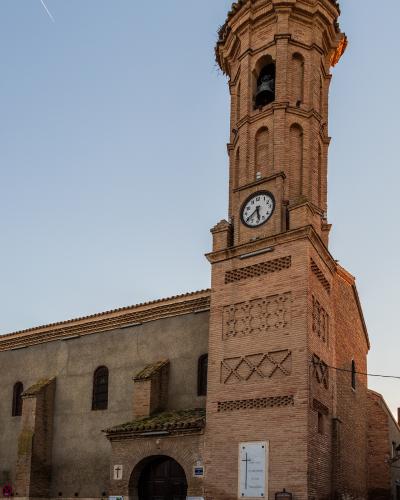 A brick church with a tall, octagonal bell tower featuring a clock and a cross, set against a clear sky.