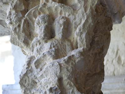 A close-up photograph of a weathered stone column capital featuring a carved relief of two figures, likely from a Romanesque cloister.