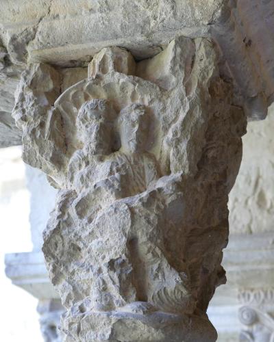 A close-up photograph of a weathered stone column capital featuring a carved relief of two figures, likely from a Romanesque cloister.