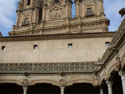 The image shows the interior courtyard (patio) of a historic building, featuring a two-story loggia with columns and ornate stonework, looking up towards a highly decorated Baroque facade with twin bell towers against a cloudy sky.