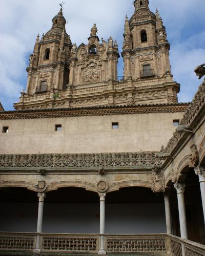 The image shows the interior courtyard (patio) of a historic building, featuring a two-story loggia with columns and ornate stonework, looking up towards a highly decorated Baroque facade with twin bell towers against a cloudy sky.