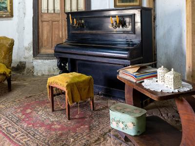 An interior room featuring a black upright piano, a yellow upholstered chair and stool, a wooden table with decorative items, and a patterned rug on a stone floor.
