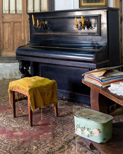 An interior room featuring a black upright piano, a yellow upholstered chair and stool, a wooden table with decorative items, and a patterned rug on a stone floor.