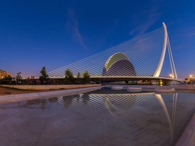 A modern white cable-stayed bridge with a tall curved pylon and a large egg-shaped building behind it, reflected in a shallow pool of water at twilight.