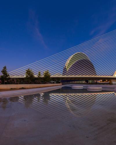 A modern white cable-stayed bridge with a tall curved pylon and a large egg-shaped building behind it, reflected in a shallow pool of water at twilight.