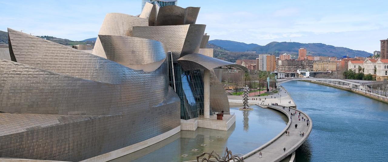 A high-angle view of the Guggenheim Museum Bilbao, featuring its distinctive titanium-clad architecture, the "Maman" spider sculpture in the foreground, and the Nervión River flowing alongside the building.