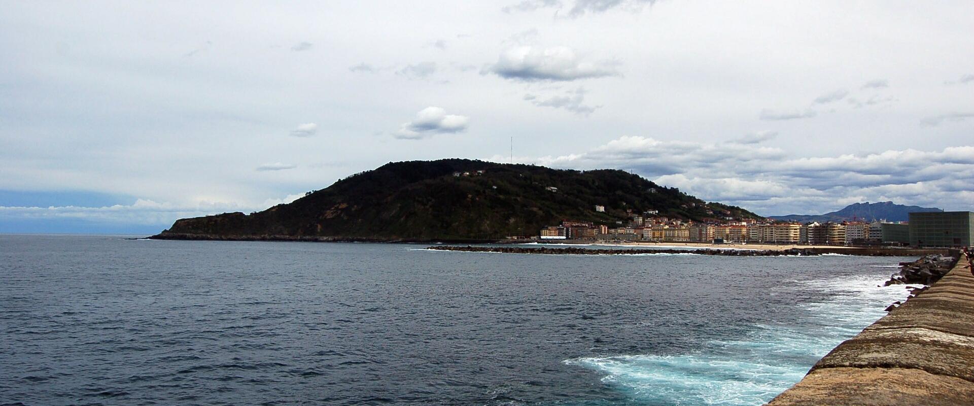 A coastal view showing a large, green, hill-shaped headland rising from the sea, with a long concrete seawall in the foreground and a row of multi-story buildings at the base of the hill.