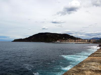 A coastal view showing a large, green, hill-shaped headland rising from the sea, with a long concrete seawall in the foreground and a row of multi-story buildings at the base of the hill.