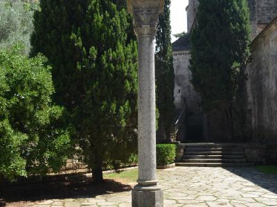 A stone wayside cross on a pedestal stands in a paved courtyard surrounded by tall cypress trees and a large Romanesque stone tower.