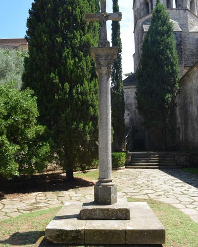 A stone wayside cross on a pedestal stands in a paved courtyard surrounded by tall cypress trees and a large Romanesque stone tower.