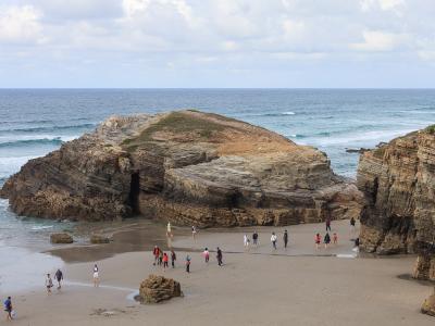 A coastal beach scene featuring large, layered rock formations and sea caves, with numerous tourists walking on the sand near the water's edge under a cloudy sky.