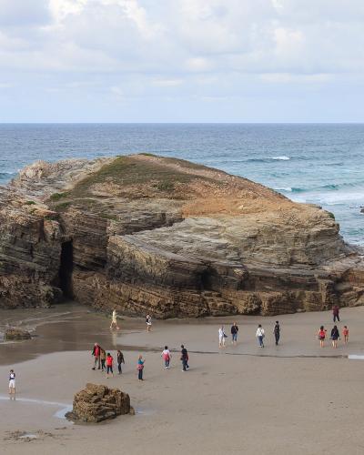 A coastal beach scene featuring large, layered rock formations and sea caves, with numerous tourists walking on the sand near the water's edge under a cloudy sky.