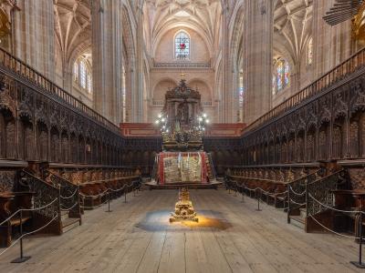 The interior of a Gothic cathedral showing ornate wooden choir stalls flanking a central aisle, with a decorated altar and stained glass windows in the background.