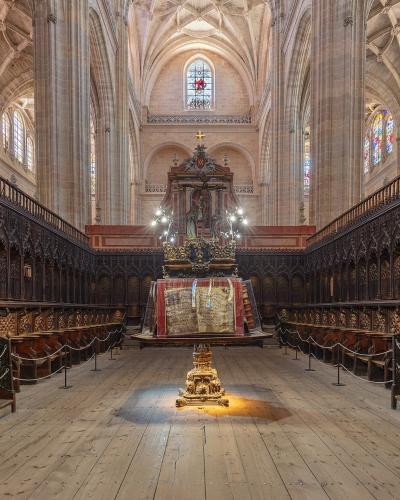 The interior of a Gothic cathedral showing ornate wooden choir stalls flanking a central aisle, with a decorated altar and stained glass windows in the background.