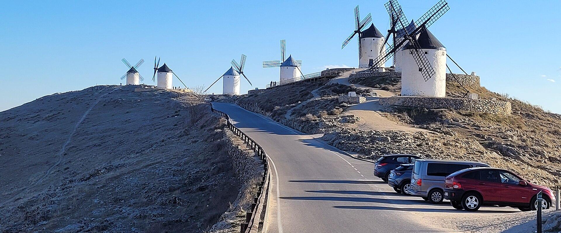 A row of white cylindrical windmills with dark conical roofs and wooden sails situated on a dry, grassy hilltop next to a paved road and parking area under a clear blue sky.