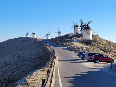 A row of white cylindrical windmills with dark conical roofs and wooden sails situated on a dry, grassy hilltop next to a paved road and parking area under a clear blue sky.