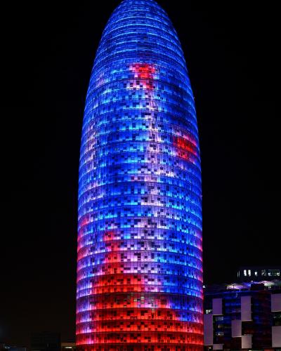A tall, bullet-shaped skyscraper illuminated at night with a red and blue pixelated light display, identified as the Torre Glòries (formerly Torre Agbar) in Barcelona.