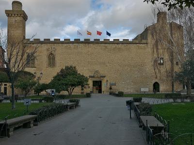 A large, fortified stone building with crenellated walls, two prominent towers, and four flags flying above the entrance, set within a landscaped park area with benches and pathways.