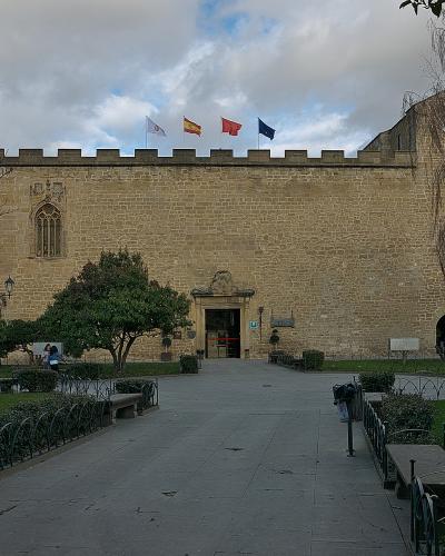 A large, fortified stone building with crenellated walls, two prominent towers, and four flags flying above the entrance, set within a landscaped park area with benches and pathways.