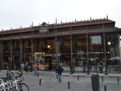 A large, ornate iron-and-glass market hall with a tiled roof, featuring prominent signage reading "MERCADO DE SAN MIGUEL" above the entrance and on the facade, with people walking outside and bicycles parked in the foreground.
