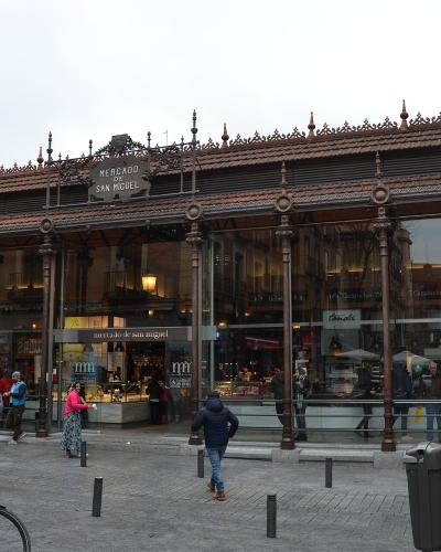 A large, ornate iron-and-glass market hall with a tiled roof, featuring prominent signage reading "MERCADO DE SAN MIGUEL" above the entrance and on the facade, with people walking outside and bicycles parked in the foreground.