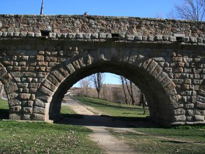 A stone Roman-style bridge with a large central arch and smaller side arches, set in a grassy area with trees and buildings visible in the background under a clear blue sky.