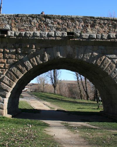 A stone Roman-style bridge with a large central arch and smaller side arches, set in a grassy area with trees and buildings visible in the background under a clear blue sky.