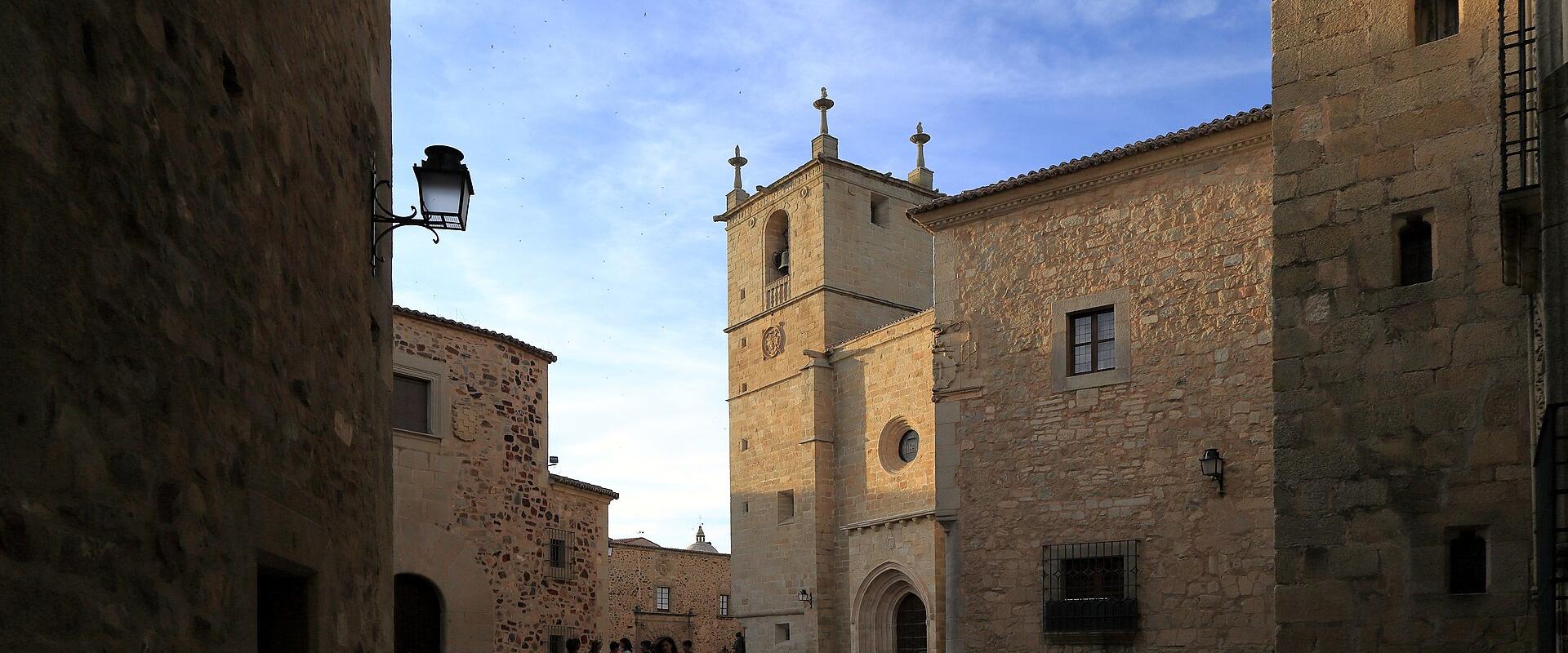 A stone plaza with a Gothic-style church tower and facade visible in the background, framed by adjacent stone buildings under a blue sky.