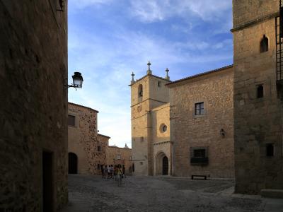 A stone plaza with a Gothic-style church tower and facade visible in the background, framed by adjacent stone buildings under a blue sky.
