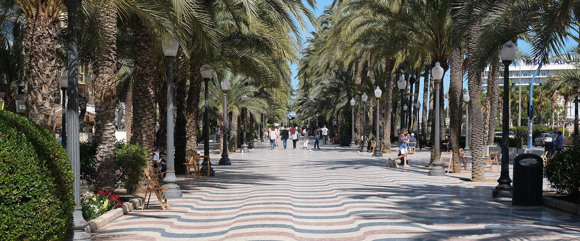 A wide pedestrian promenade lined with tall palm trees and distinctive black lampposts, featuring a unique wavy-patterned pavement in red, white, and blue.