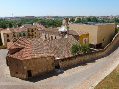 A high-angle view of a historic stone building with a terracotta tiled roof, adjacent to a modern beige structure, with the word "MUSEO" visible on a low stone wall in the foreground and a cityscape in the background.