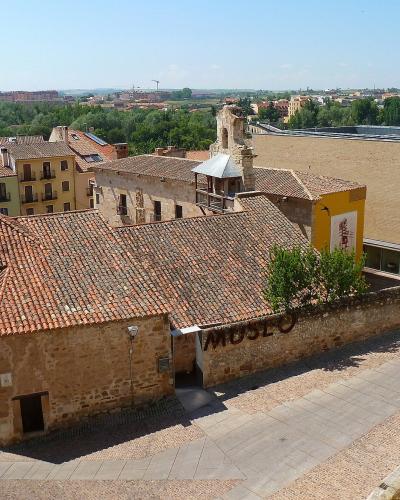 A high-angle view of a historic stone building with a terracotta tiled roof, adjacent to a modern beige structure, with the word "MUSEO" visible on a low stone wall in the foreground and a cityscape in the background.