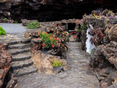 A landscaped volcanic rock garden with stone pathways, steps, and planted flowers leading toward a dark cave entrance, featuring distinctive white-washed architectural elements integrated into the black lava rock.