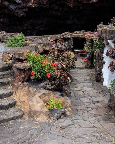 A landscaped volcanic rock garden with stone pathways, steps, and planted flowers leading toward a dark cave entrance, featuring distinctive white-washed architectural elements integrated into the black lava rock.