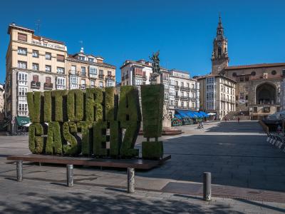 A large, green topiary sign spelling "VITORIA GASTEIZ" stands in a paved public square, with historic buildings and a church tower in the background.