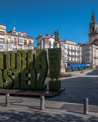 A large, green topiary sign spelling "VITORIA GASTEIZ" stands in a paved public square, with historic buildings and a church tower in the background.