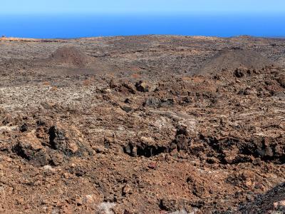 A barren volcanic landscape with reddish-brown cinder cones and dark lava fields under a clear blue sky, with the ocean visible on the horizon.