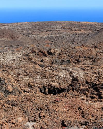 A barren volcanic landscape with reddish-brown cinder cones and dark lava fields under a clear blue sky, with the ocean visible on the horizon.