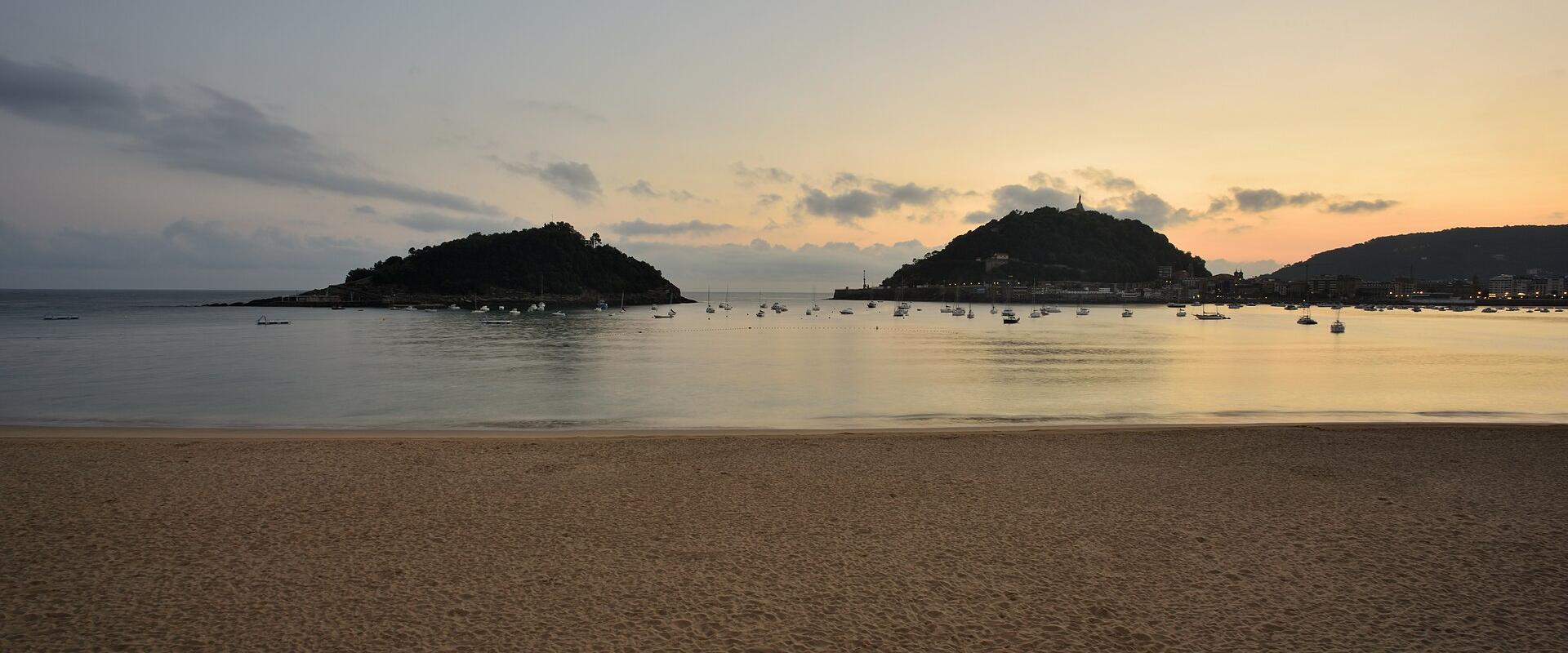 A wide sandy beach at dusk or dawn with calm water, numerous anchored boats, and two prominent hills in the background, one of which features a tower on its summit.