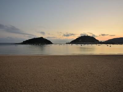 A wide sandy beach at dusk or dawn with calm water, numerous anchored boats, and two prominent hills in the background, one of which features a tower on its summit.