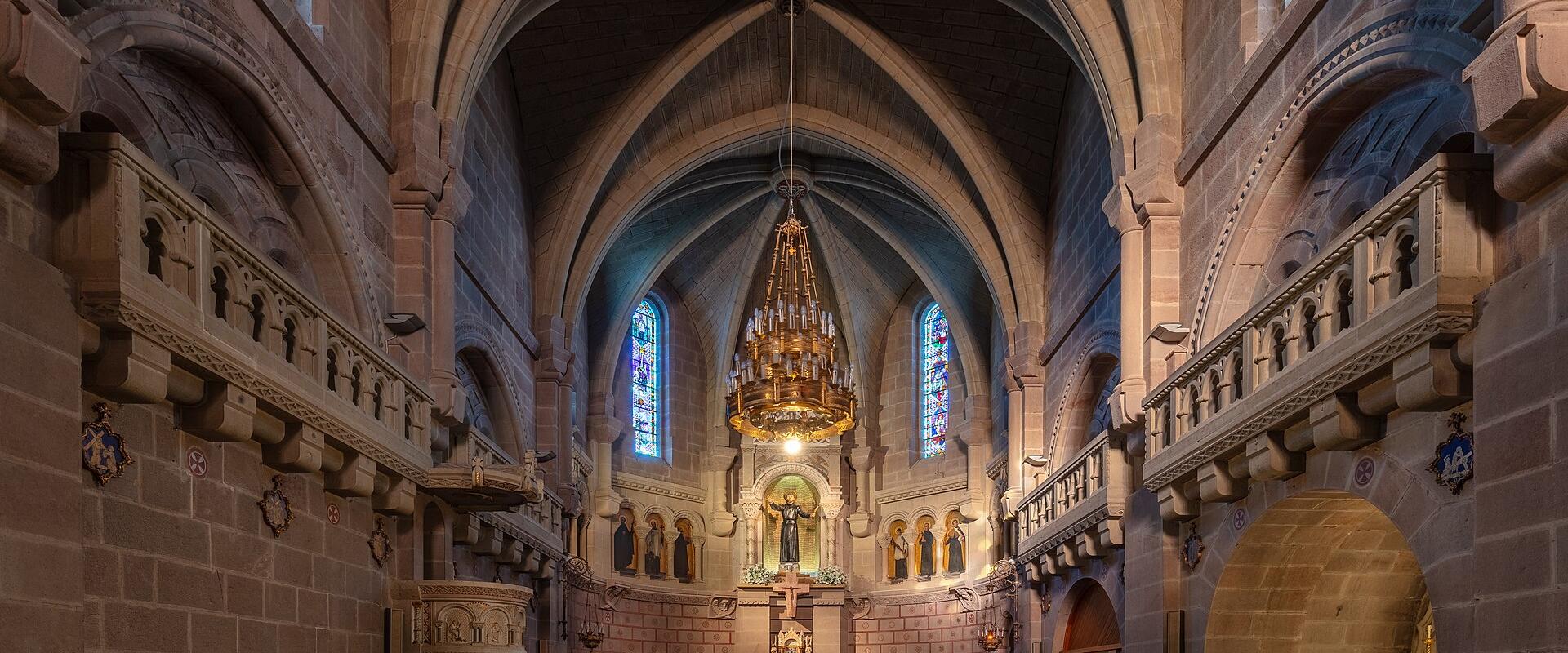 The interior of a stone church with vaulted ceilings, wooden pews, a large chandelier, and stained glass windows, featuring a balcony with coats of arms on the side walls.