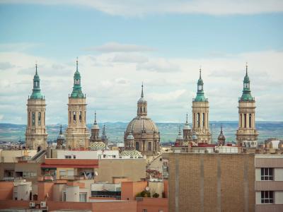 A cityscape view of the Basilica del Pilar in Zaragoza, Spain, featuring its distinctive towers and dome against a cloudy sky, with urban rooftops in the foreground.