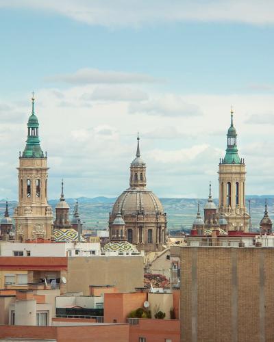 A cityscape view of the Basilica del Pilar in Zaragoza, Spain, featuring its distinctive towers and dome against a cloudy sky, with urban rooftops in the foreground.